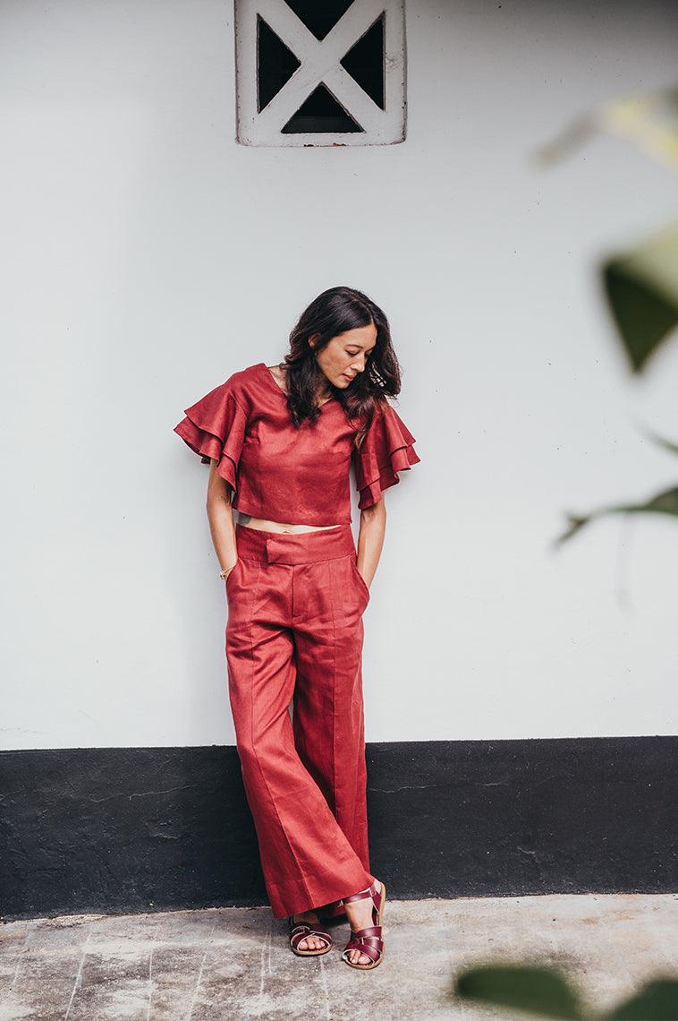 Adult with brown hair wearing a red jumper. Leaning against a white wall with a black stripe at the bottom. The adult is crossing their feet wearing our claret salt water originals.