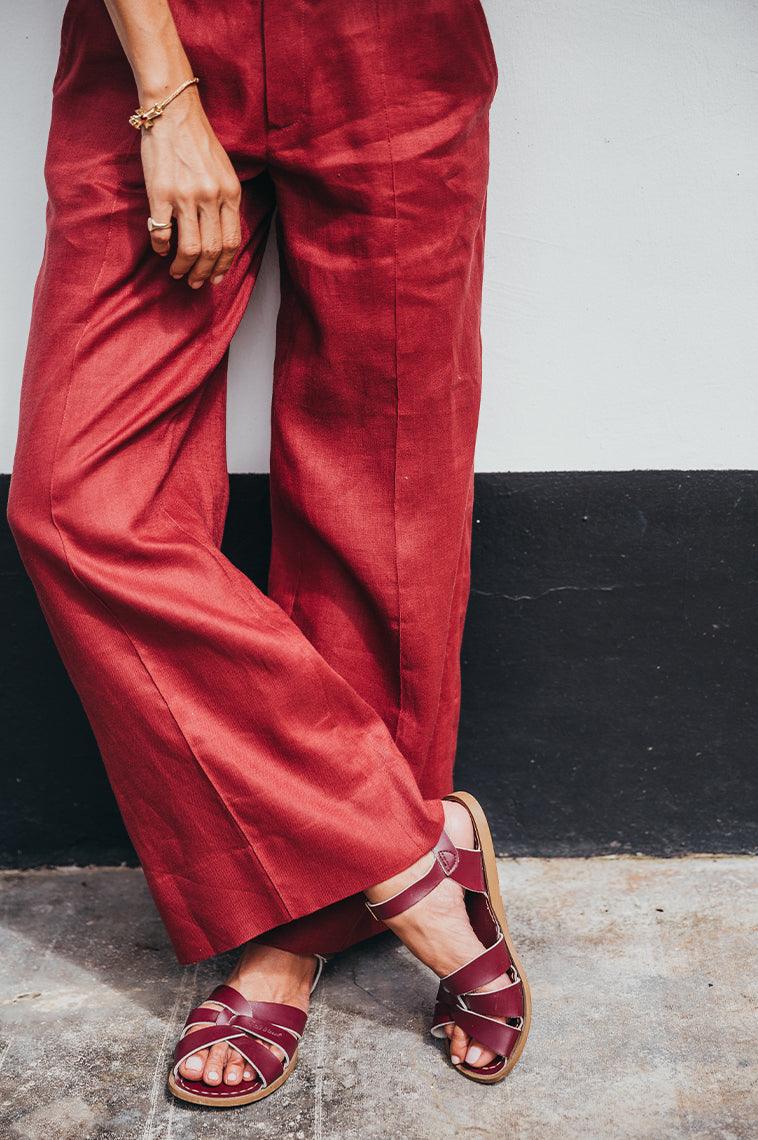 An close up photo of an adults wearing red pants, leaning against a white wall with a black  stripe at the bottom. The adult is crossing their feet wearing our claret originals.